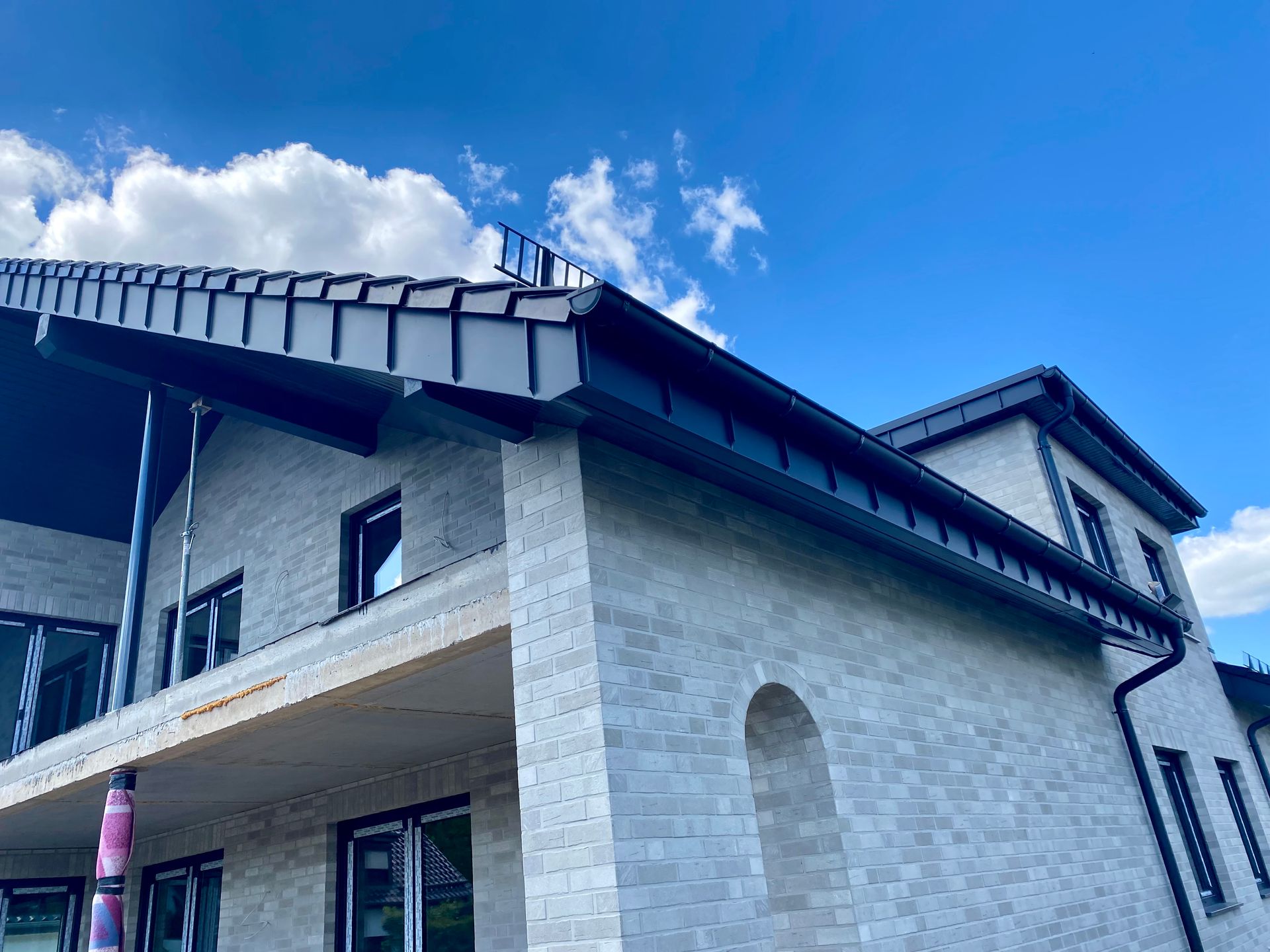 Ein teilweise errichtetes Haus mit hellen Steinmauern, dunklem Metalldach und blauem Himmel mit vereinzelten Wolken.