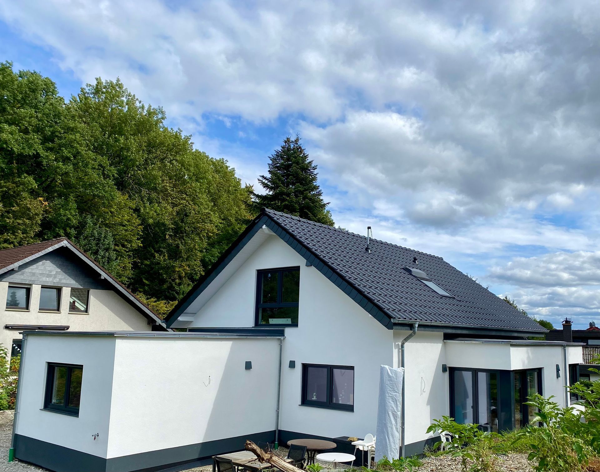 Ein modernes weißes Haus mit dunklem Giebeldach und Flachdachanbauten vor blauem, bewölktem Himmel und grünen Bäumen.
