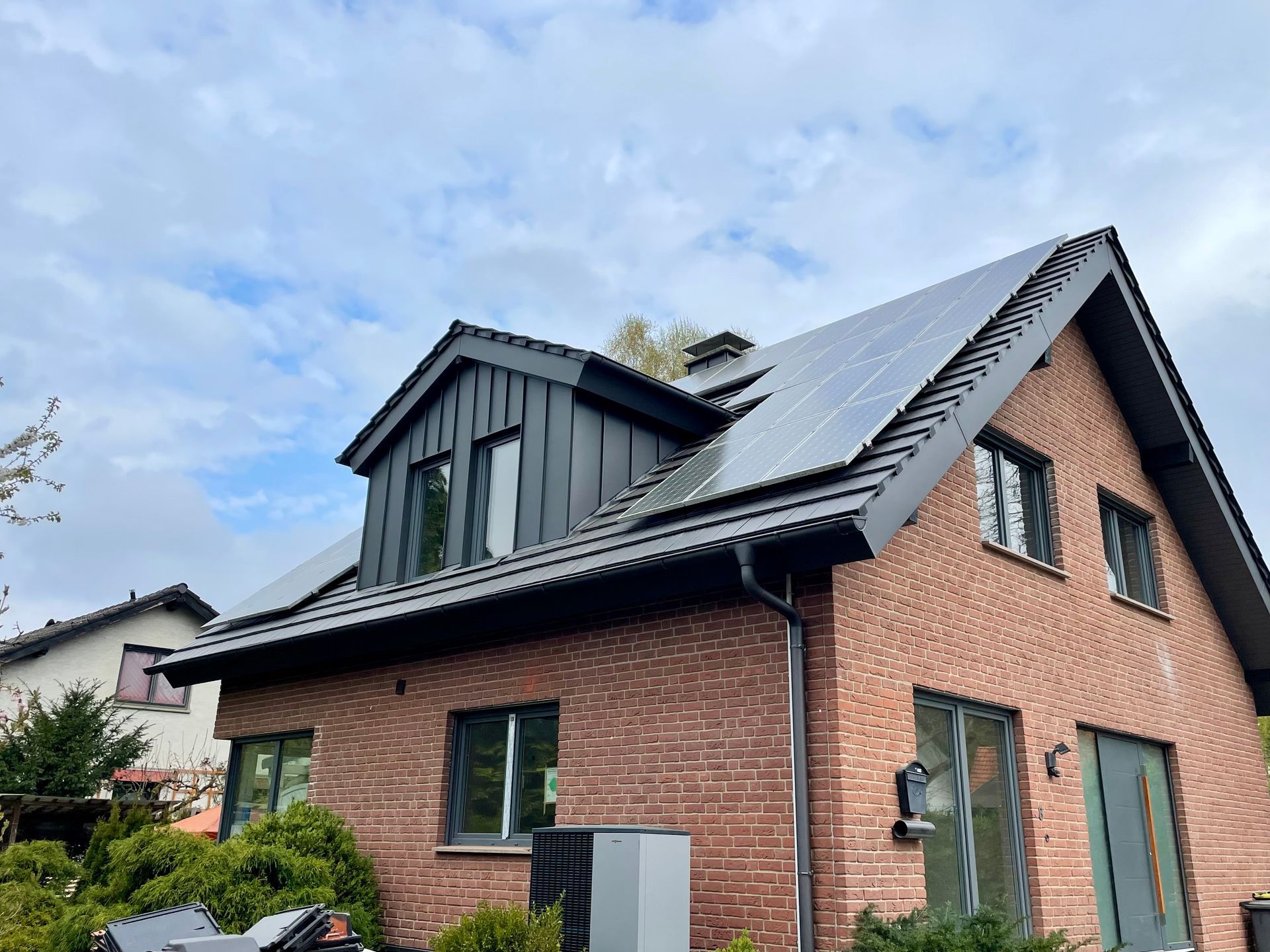 Ein Haus aus roten Ziegelsteinen mit einer dunkelmetallverkleideten Dachgaube und Solarpaneelen auf dem Dach