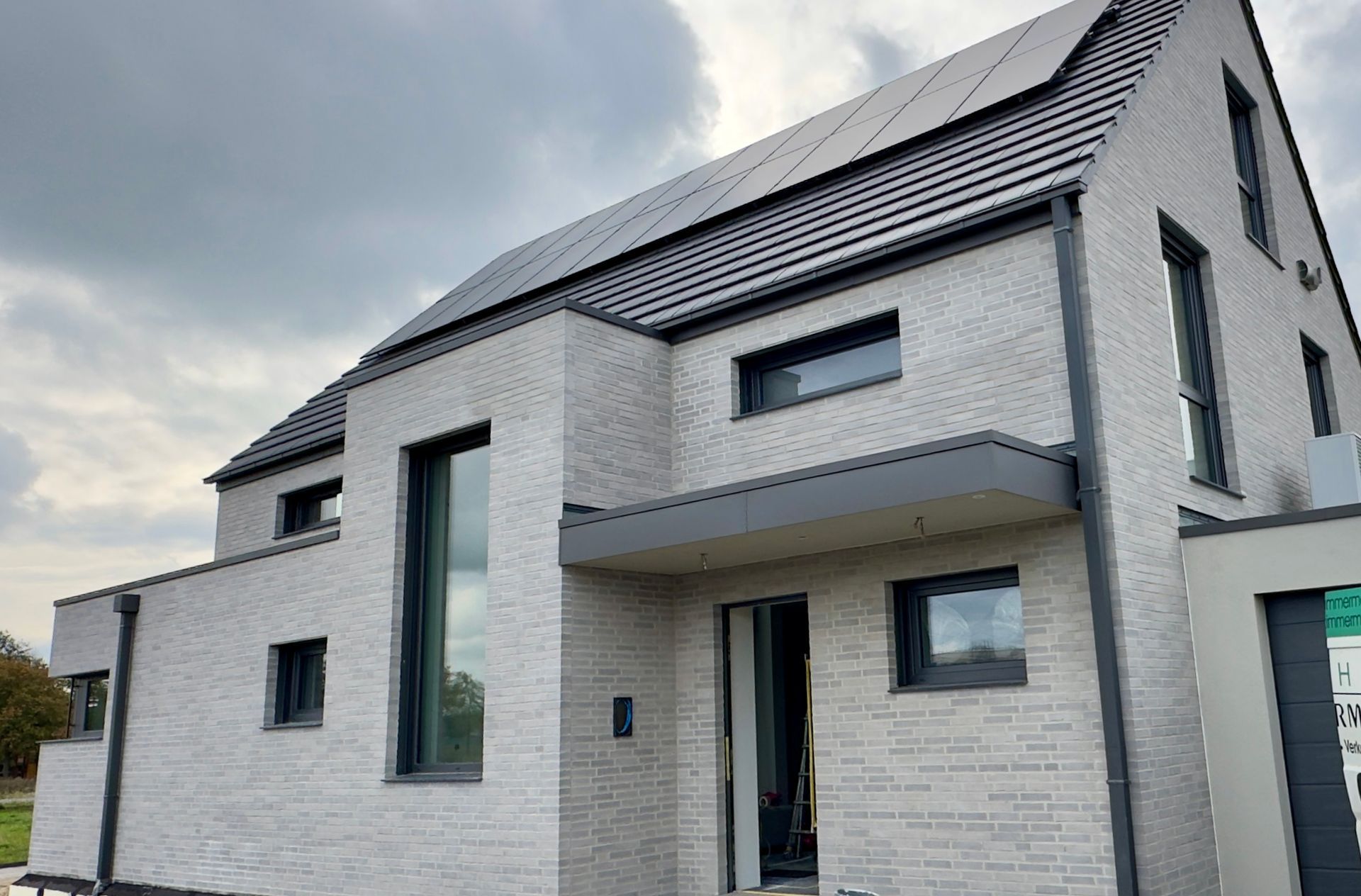 Ein modernes zweistöckiges Haus mit hellgrauer Backsteinfassade, dunklen Fensterrahmen und Solarpaneelen auf dem Dach.