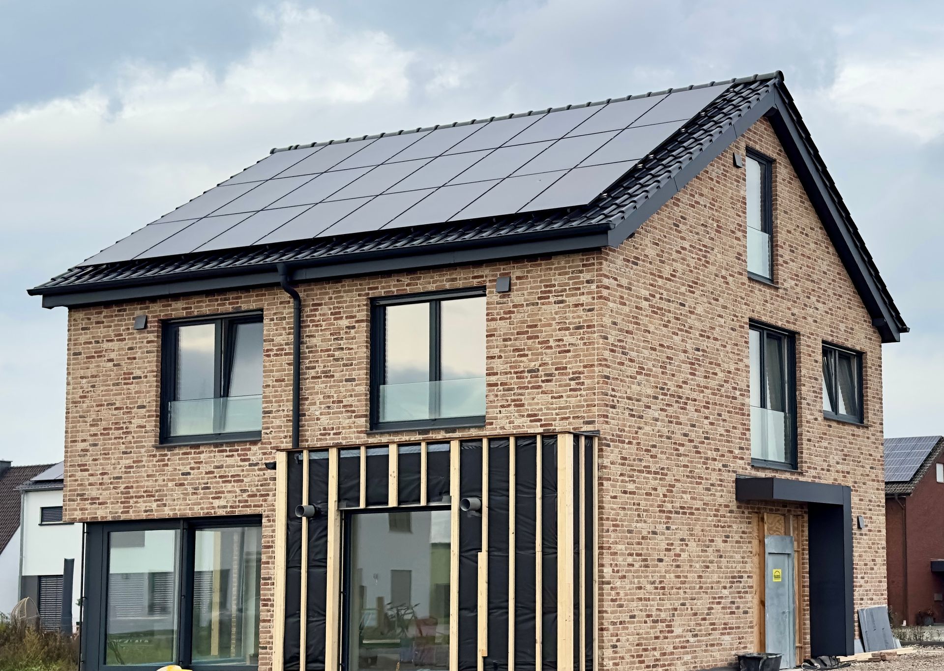 Ein modernes zweistöckiges Backsteinhaus mit Solardach und laufenden Bauarbeiten an der Außenwand an einem bewölkten Tag.