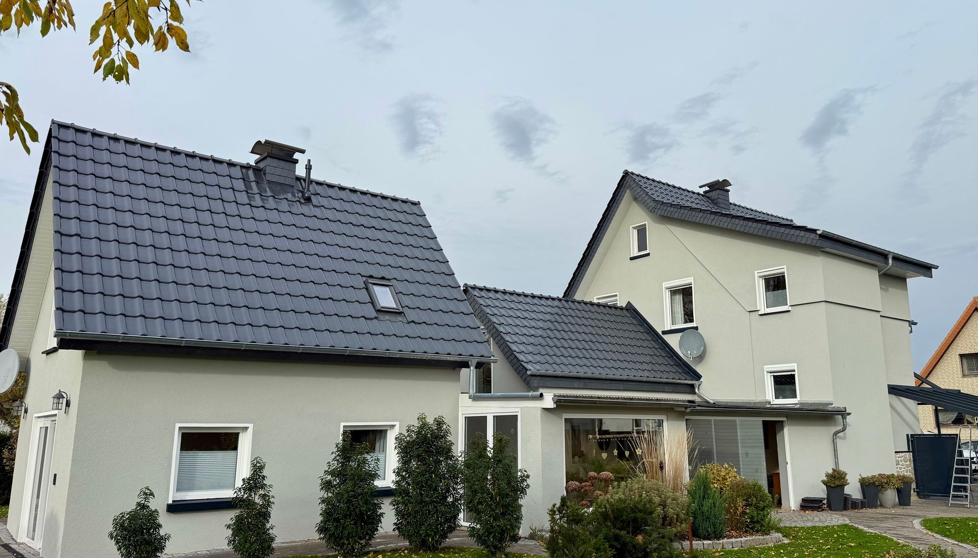 Ein zweistöckiges, hellgraues Wohnhaus mit dunklem Ziegeldach und kleinen Sträuchern im Vorgarten.