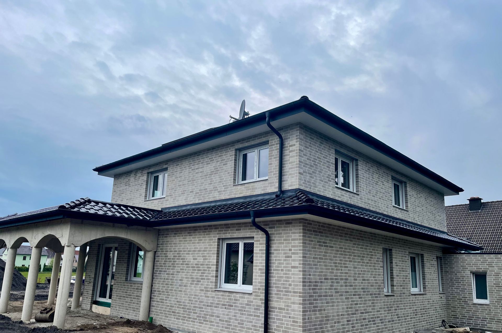 Ein zweistöckiges Haus aus hellen Ziegelsteinen mit schwarzem Dach und überdachter Veranda mit Säulen unter bewölktem Himmel.