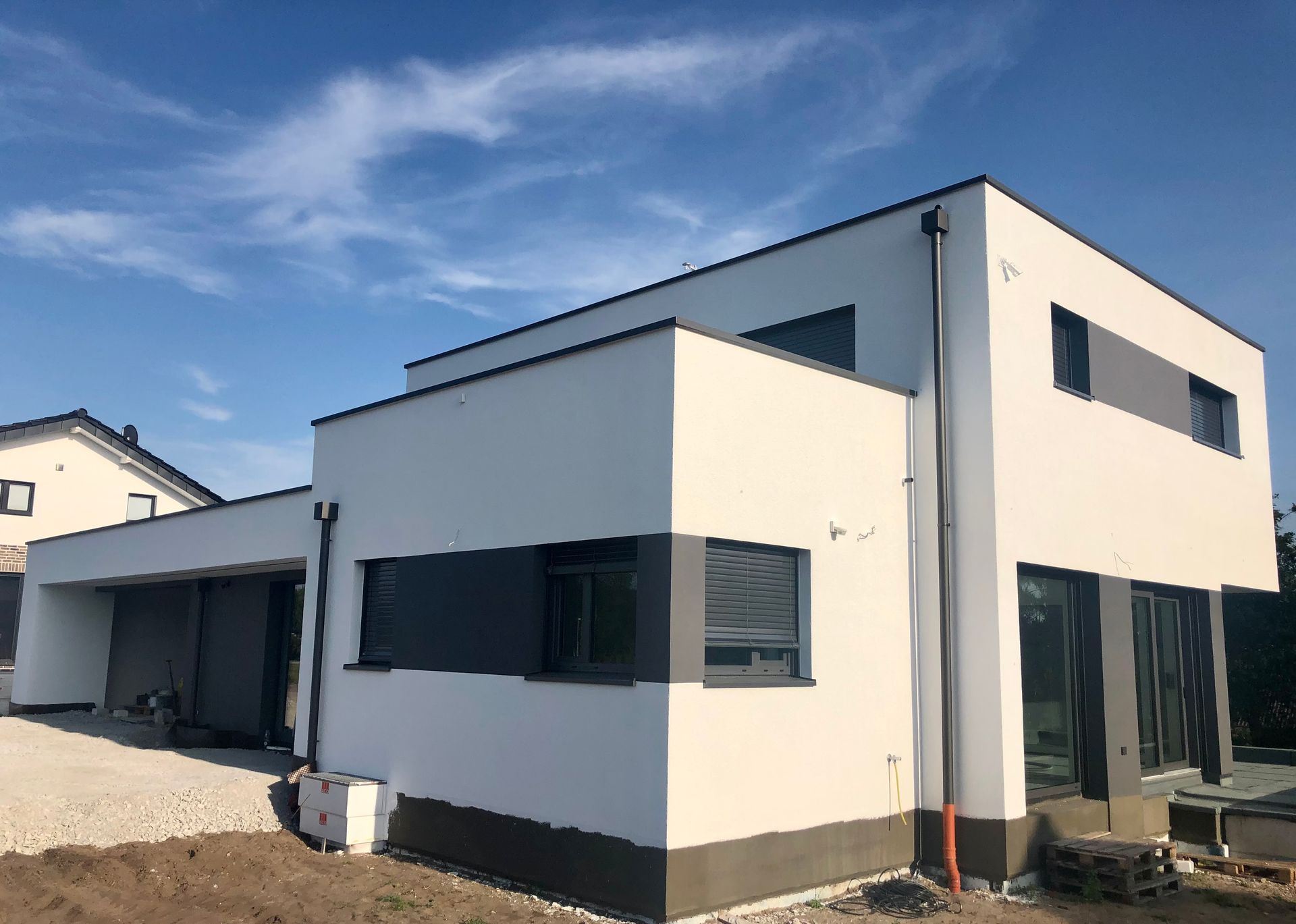 Ein modernes zweistöckiges Haus mit Flachdach, weißen Wänden und dunkelgrauen Akzentpaneelen vor einem klaren blauen Himmel.