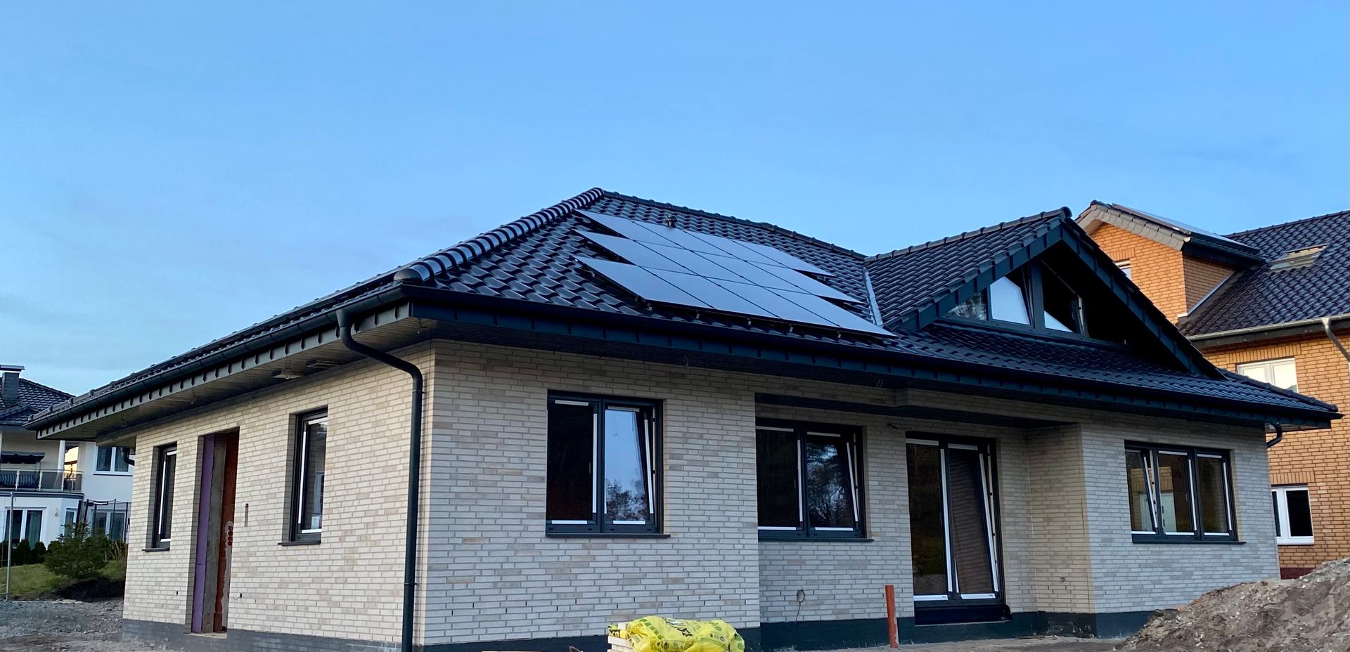 Ein eingeschossiges, helles Backsteinhaus mit dunklem Ziegeldach und Solarpaneelen unter klarem, blauem Himmel.
