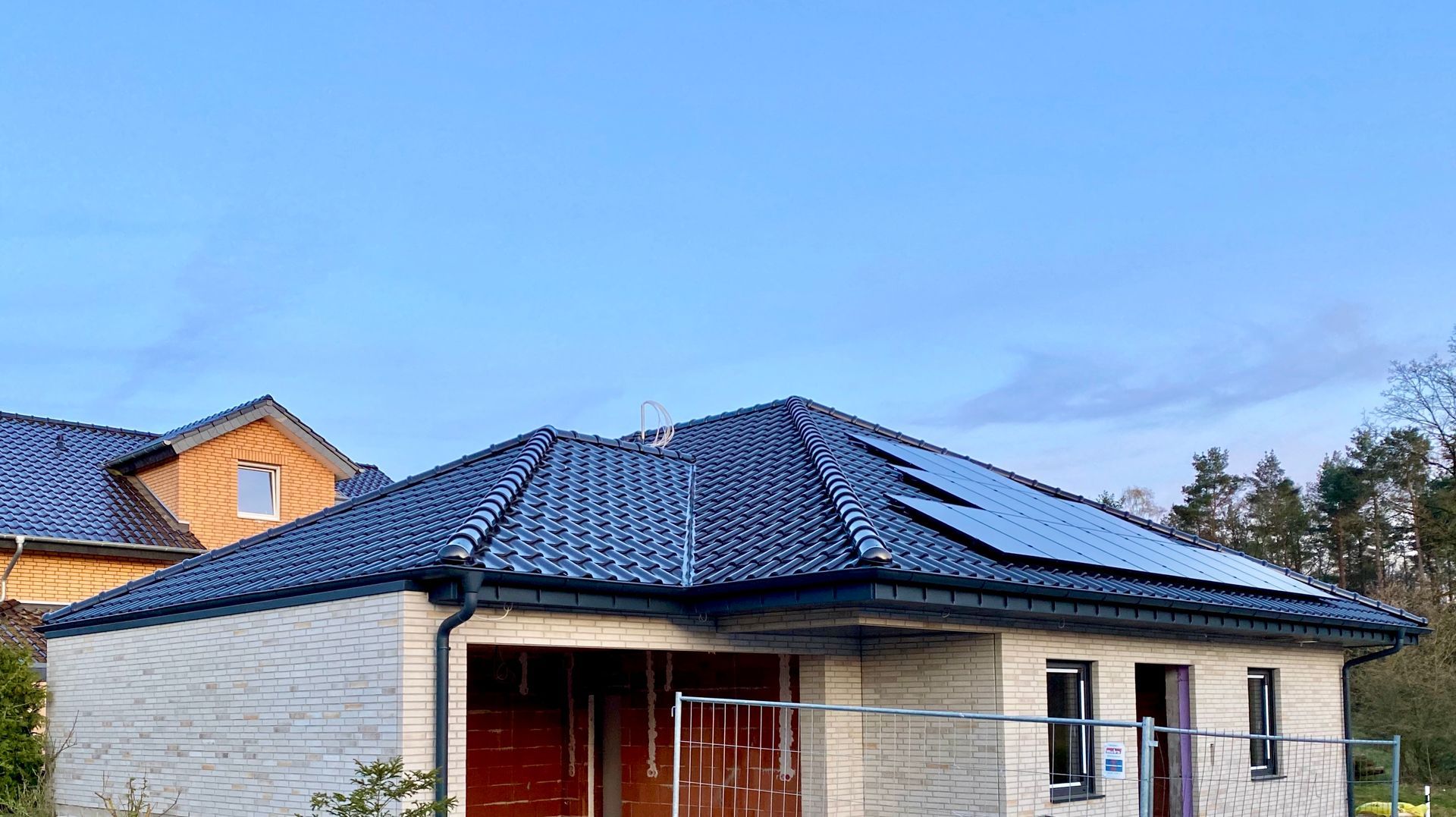 Ein eingeschossiges Haus mit blauem Ziegeldach, Solarpaneelen und unfertigen Außenwänden unter klarem, blauem Himmel.