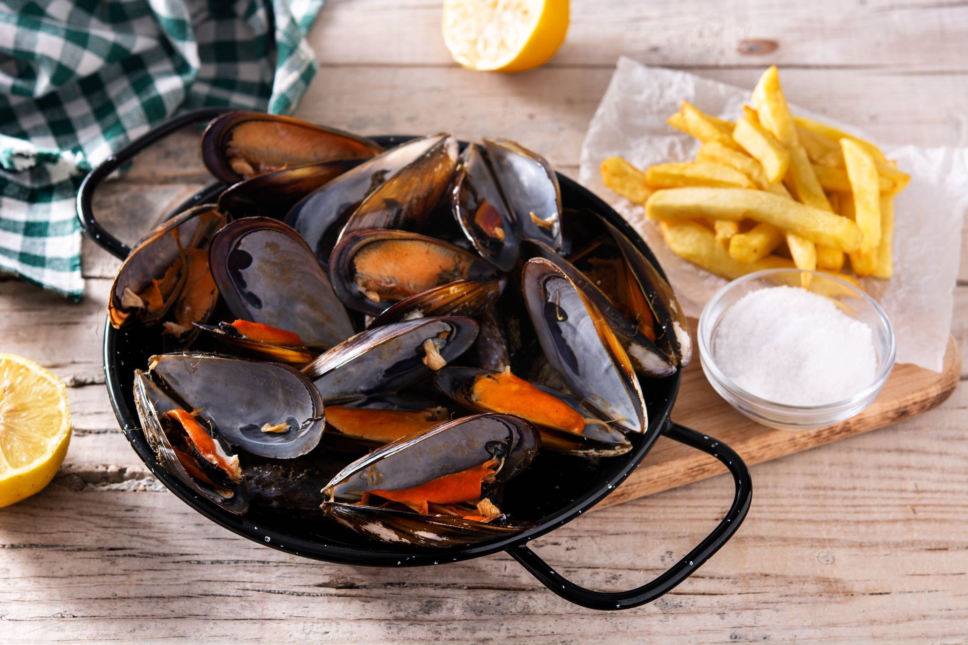 Moules cuites à la vapeur dans une poêle noire, accompagnées de frites, de quartiers de citron et de sel, sur une table en bois.