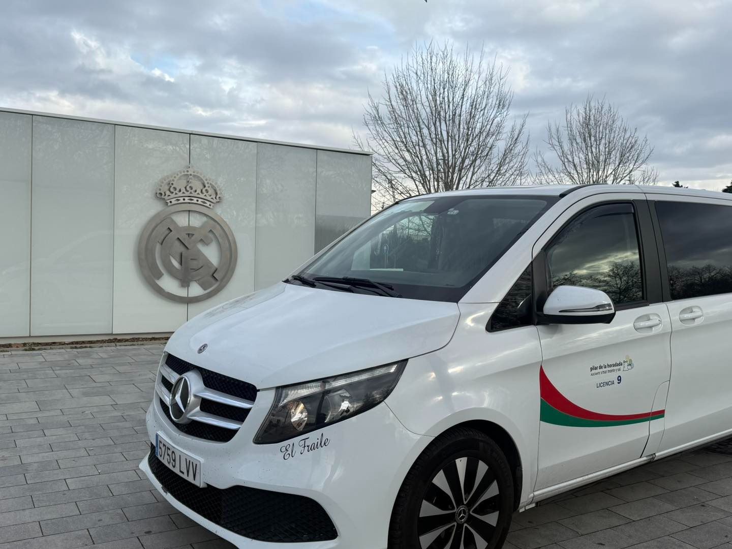 Una furgoneta Mercedes blanca estacionada frente a un gran emblema plateado del Real Madrid. La furgoneta tiene una franja verde y roja.