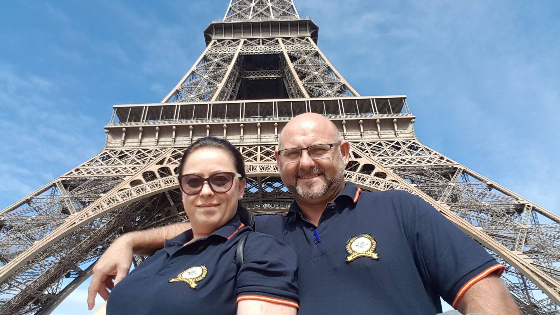 Una pareja posando frente a la Torre Eiffel en París, Francia. Llevan camisetas azules y gafas de sol a juego.