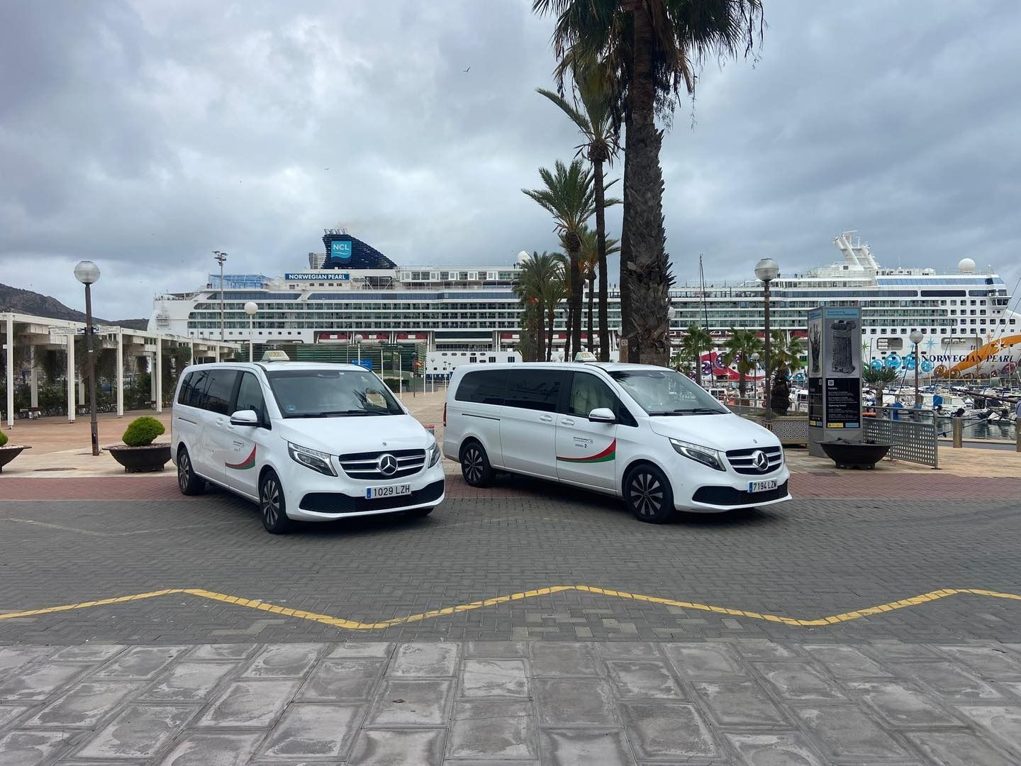 Dos furgonetas Mercedes-Benz blancas con distintivos de taxi estacionadas frente a un crucero. Día nublado en un puerto.