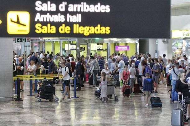 Una sala de llegadas del aeropuerto abarrotada de gente esperando, equipaje y carteles direccionales en español e inglés.