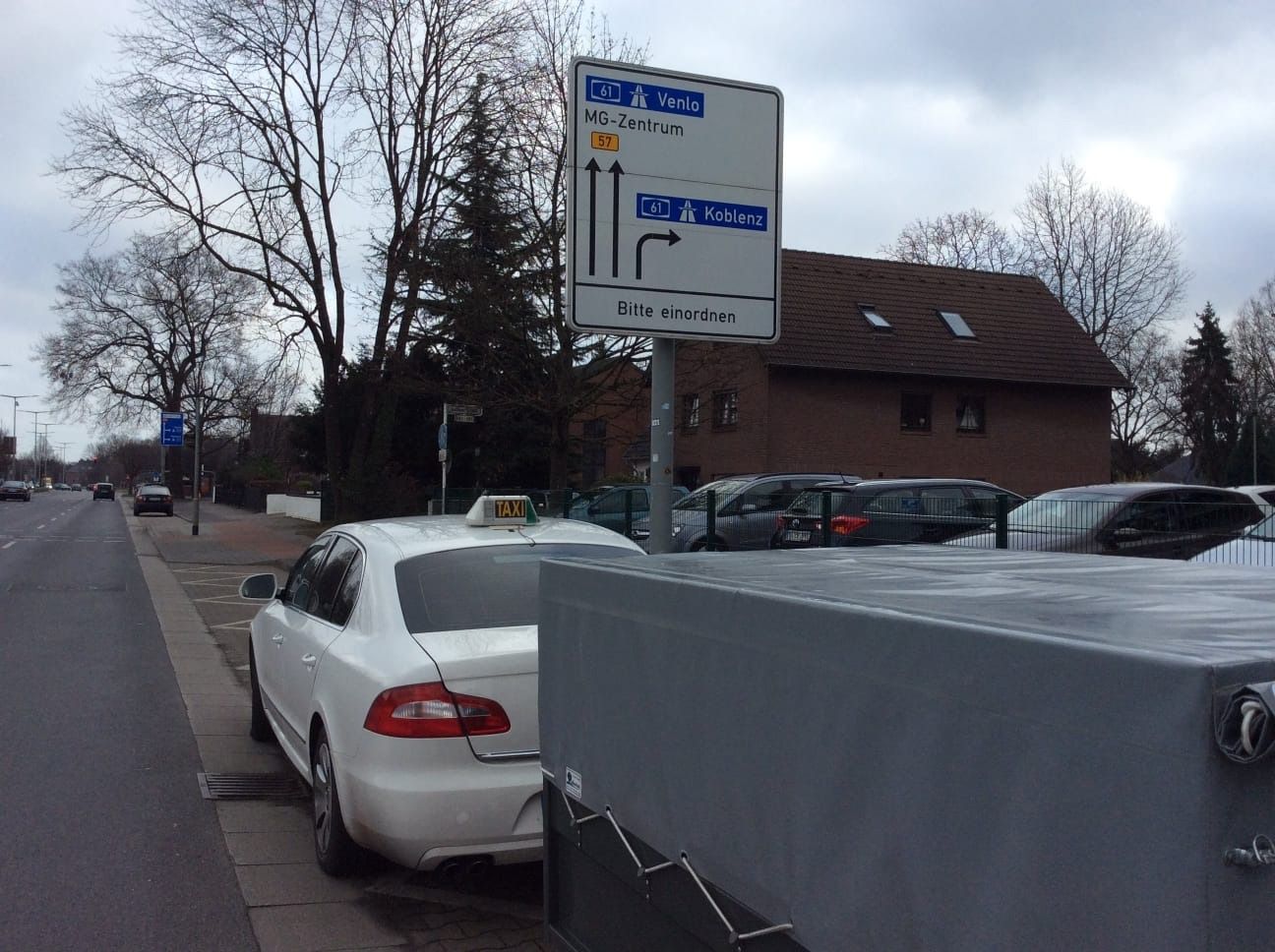 Un taxi blanco estacionado junto a la acera con una señal de tráfico. En primer plano, se ve una caja gris, con un edificio y coches aparcados detrás del taxi.