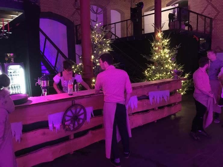 Eine Gruppe Leute steht um eine Holzbar herum , im Hintergrund ist ein Weihnachtsbaum zu sehen.