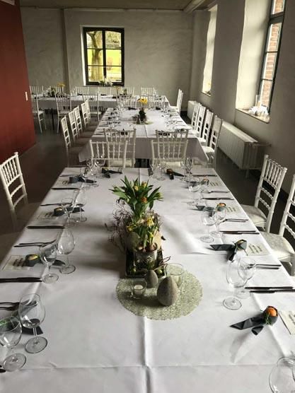 Ein langer Tisch mit einer weißen Tischdecke und einer Vase mit Blumen darauf.