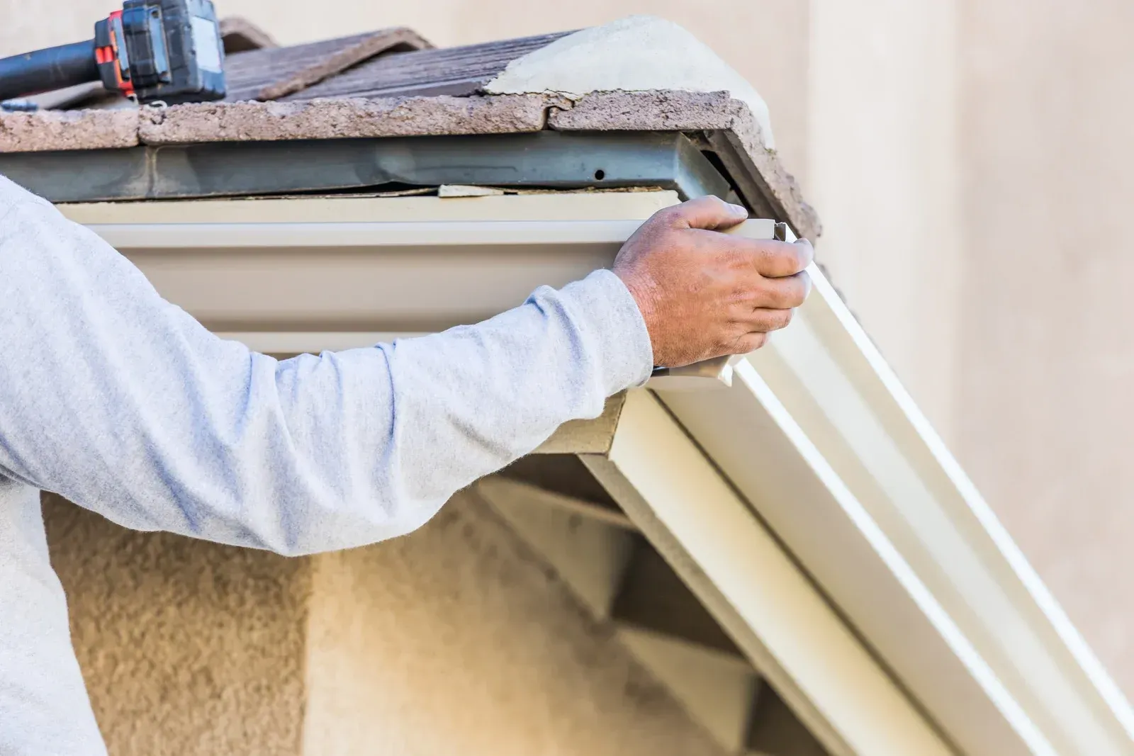 Worker installing white gutter on house roof with a power drill