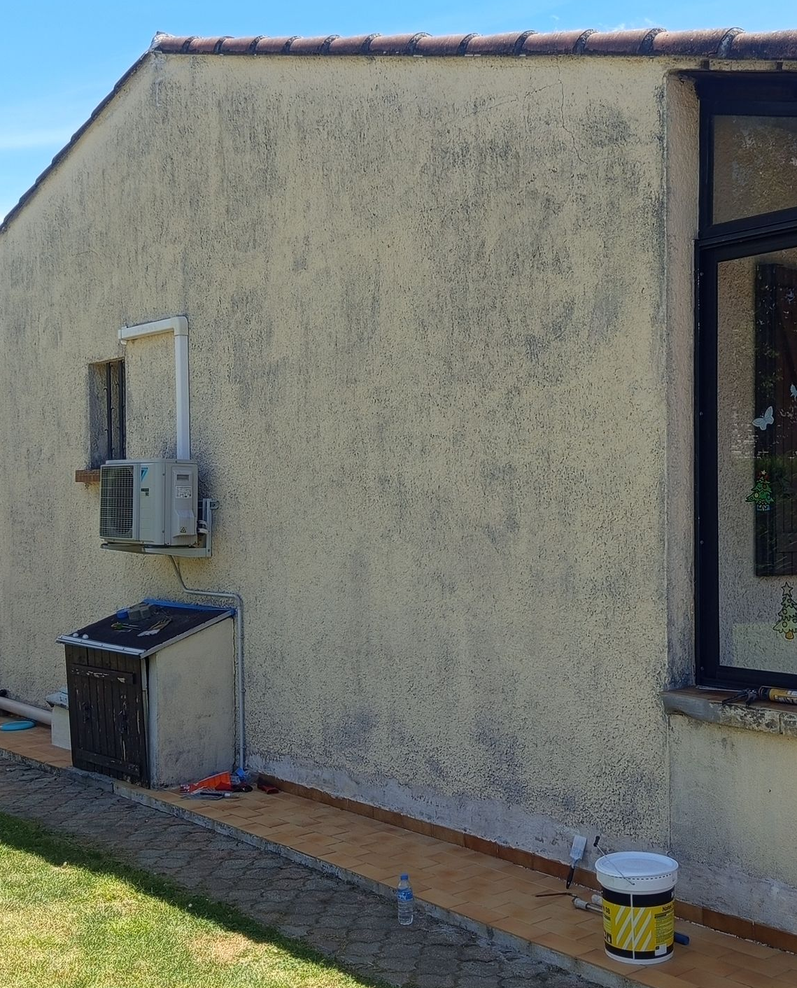 Côté d'une maison en stuc avec un climatiseur de fenêtre, une poubelle et un seau jaune le long du mur.