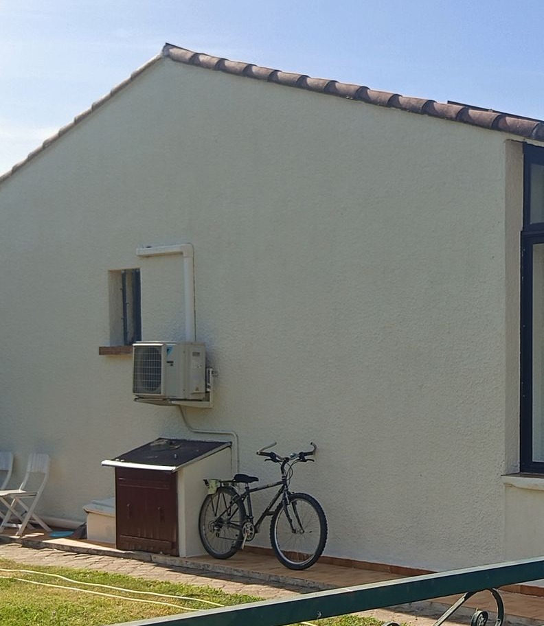 Côté d'une maison beige avec un vélo et un petit coffre de rangement contre le mur, sous un toit de tuiles.