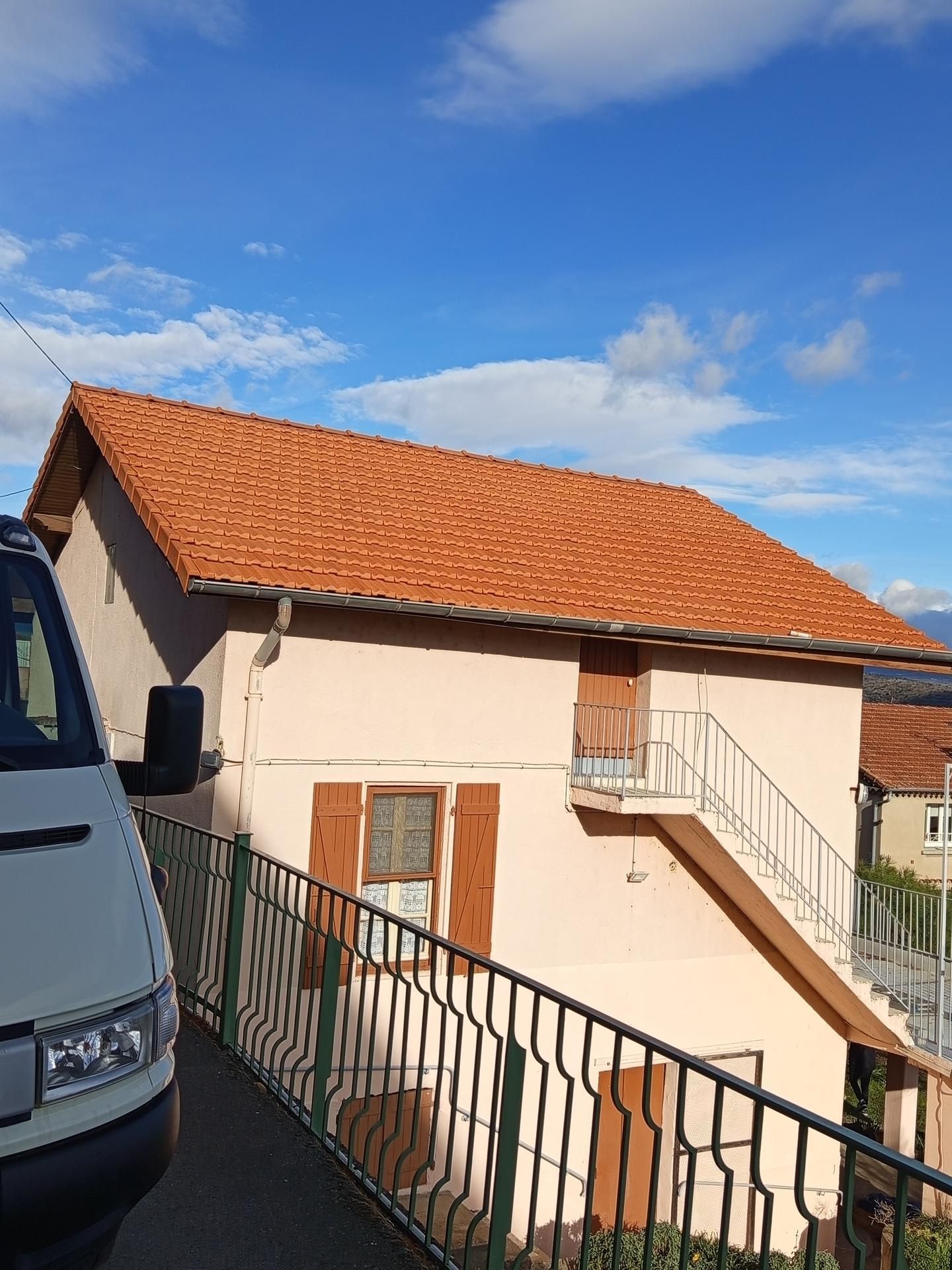 Extérieur d'une maison couleur pêche avec toit de tuiles rouges, balustrade verte et ciel bleu, partiellement masqué par une camionnette