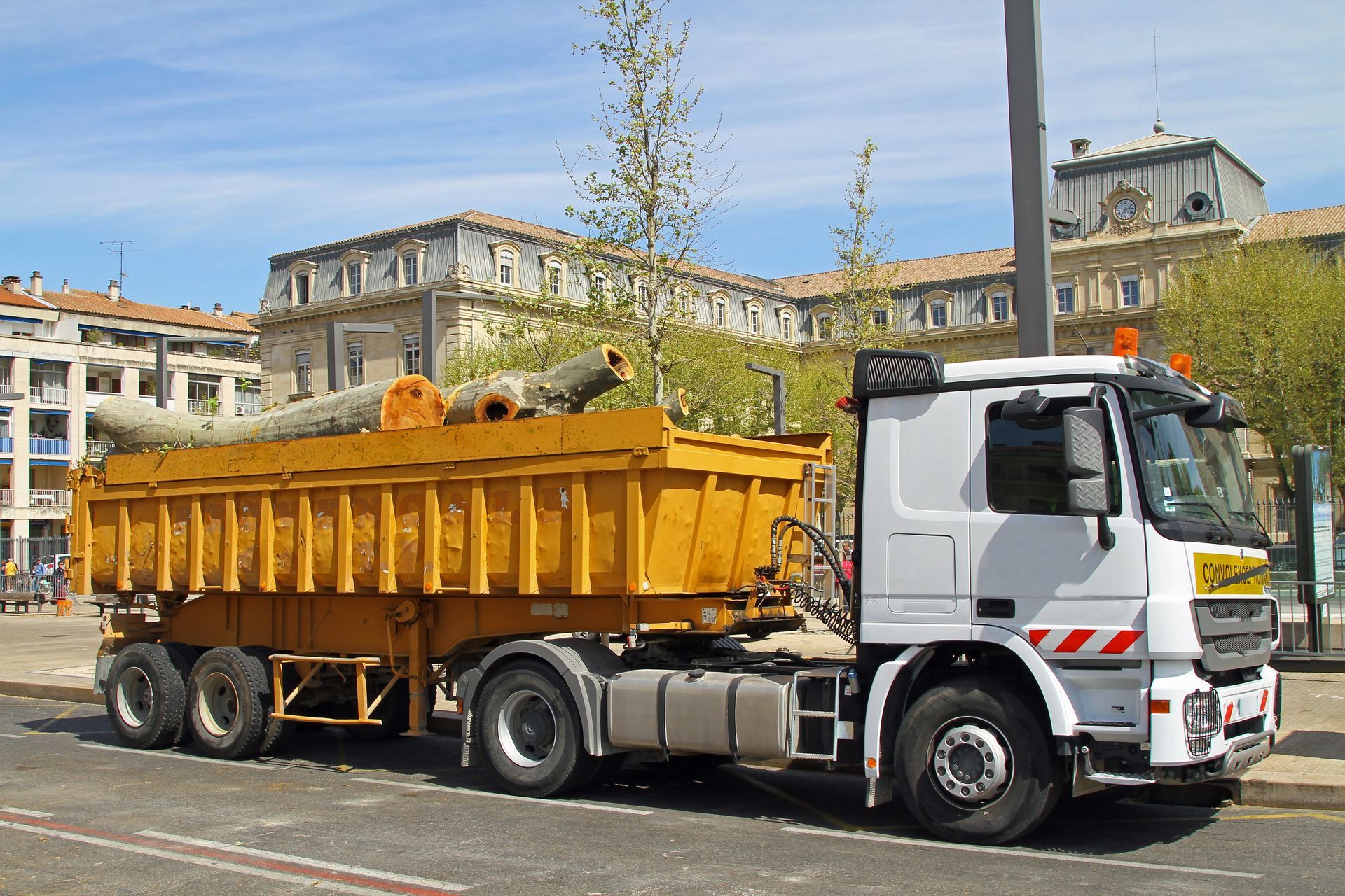 Un camion blanc tractant une benne basculante jaune circule dans une rue de la ville sous un ciel bleu.