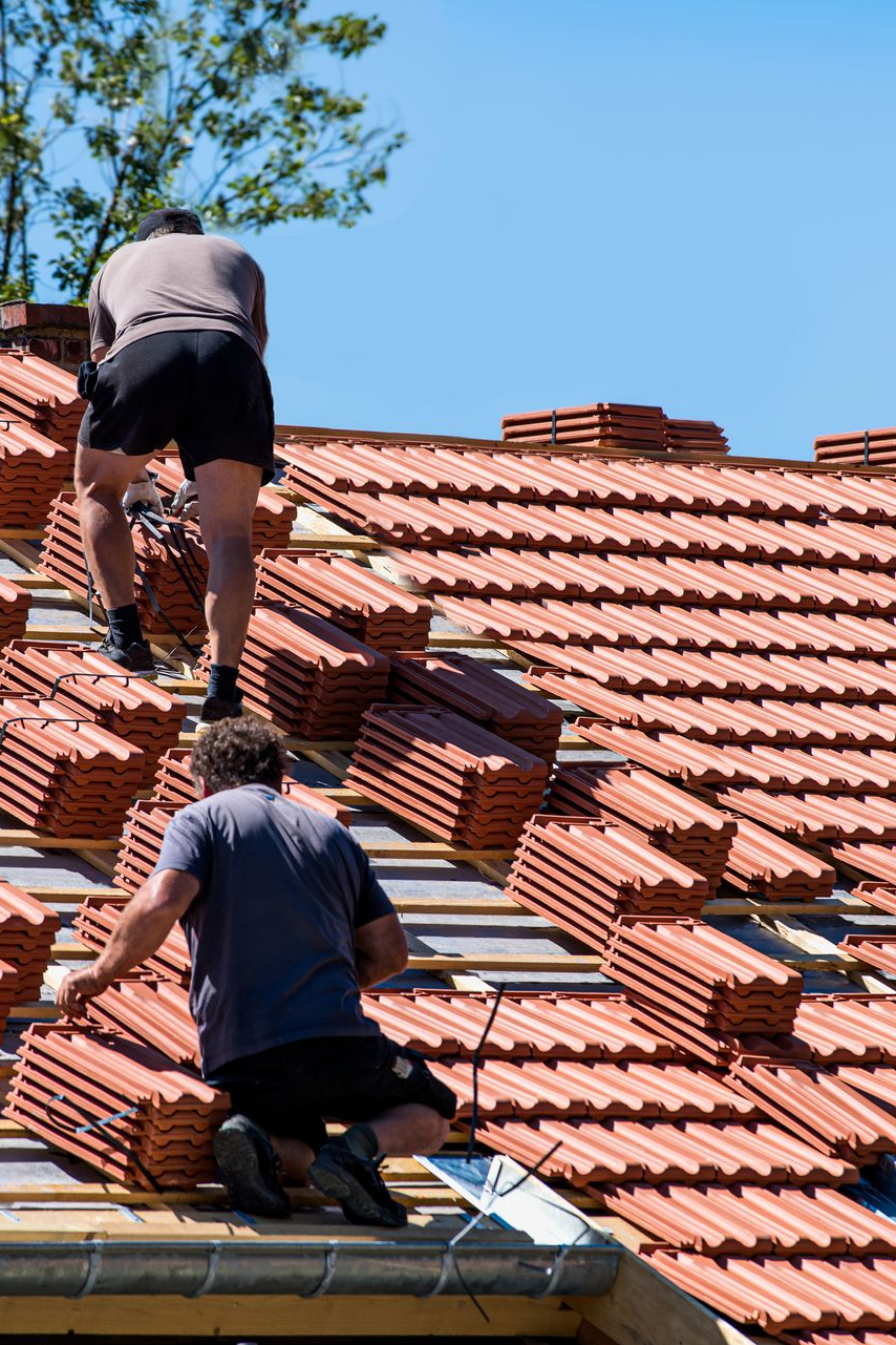 Deux couvreurs installent des tuiles en terre cuite sur un toit pentu de tuiles rouges sous un ciel bleu.