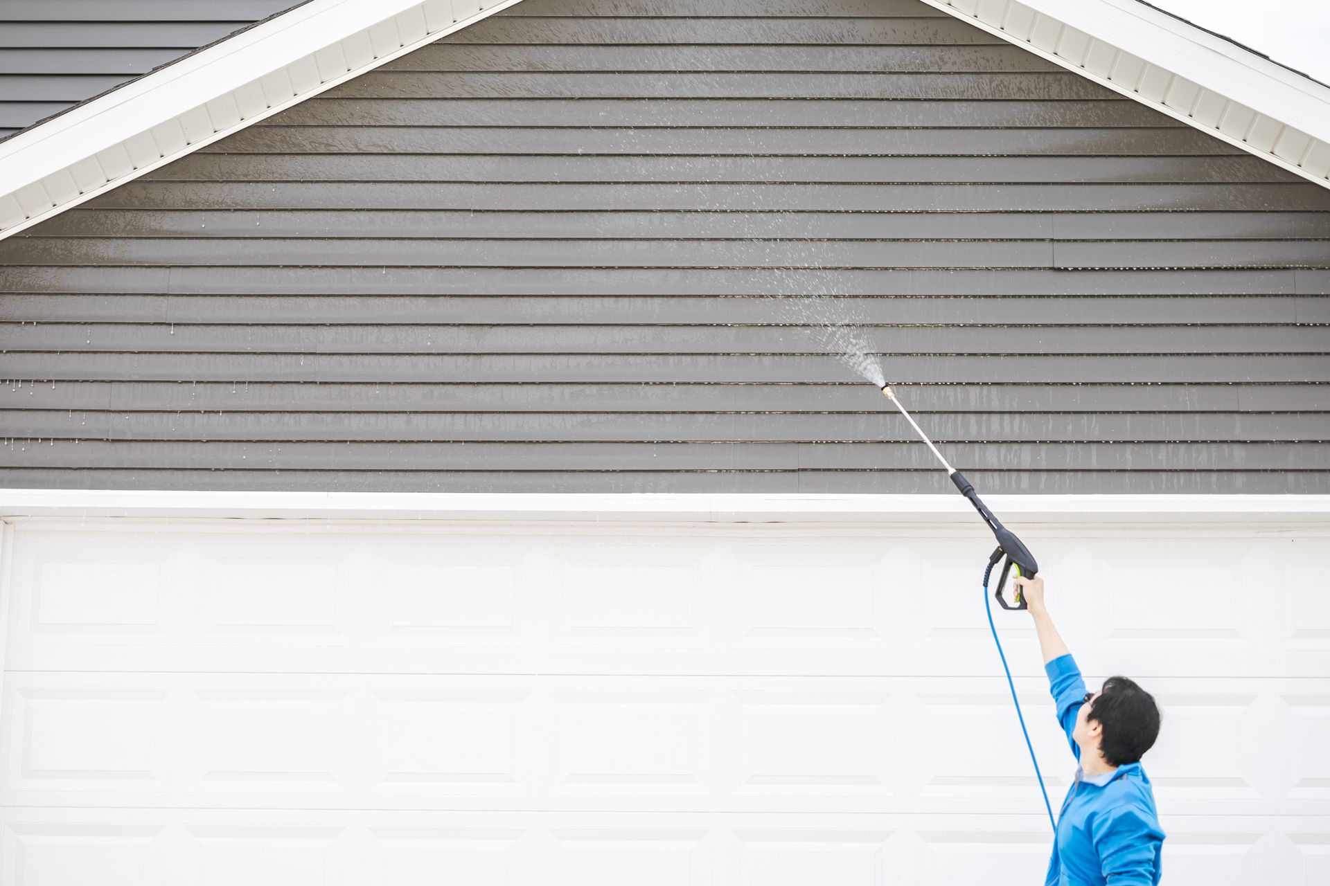 Une personne nettoie à haute pression le revêtement gris d'une maison et une porte de garage blanche à l'aide d'une longue lance.