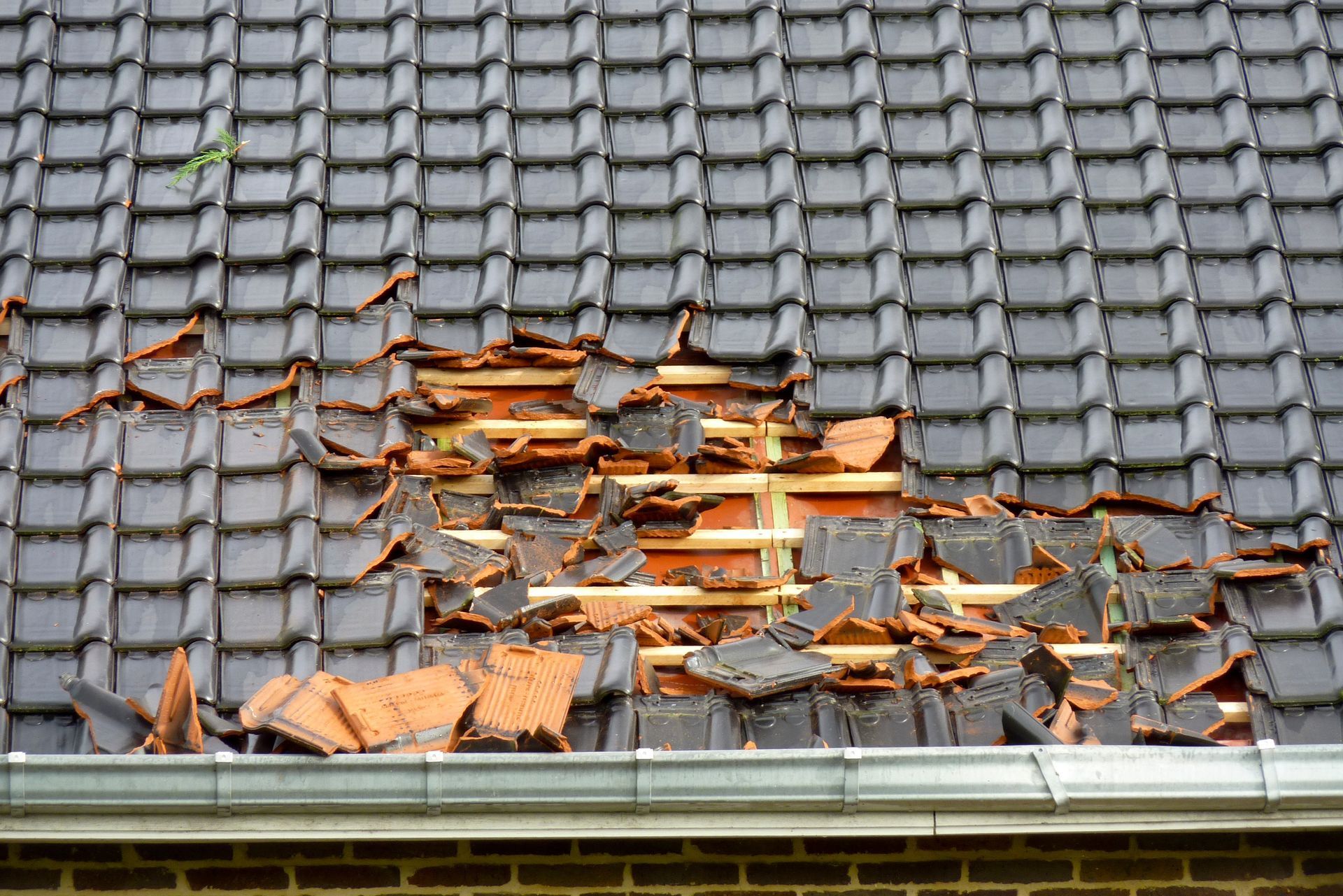 Toiture grise endommagée, avec des tuiles manquantes et une sous-couche en bois apparente.