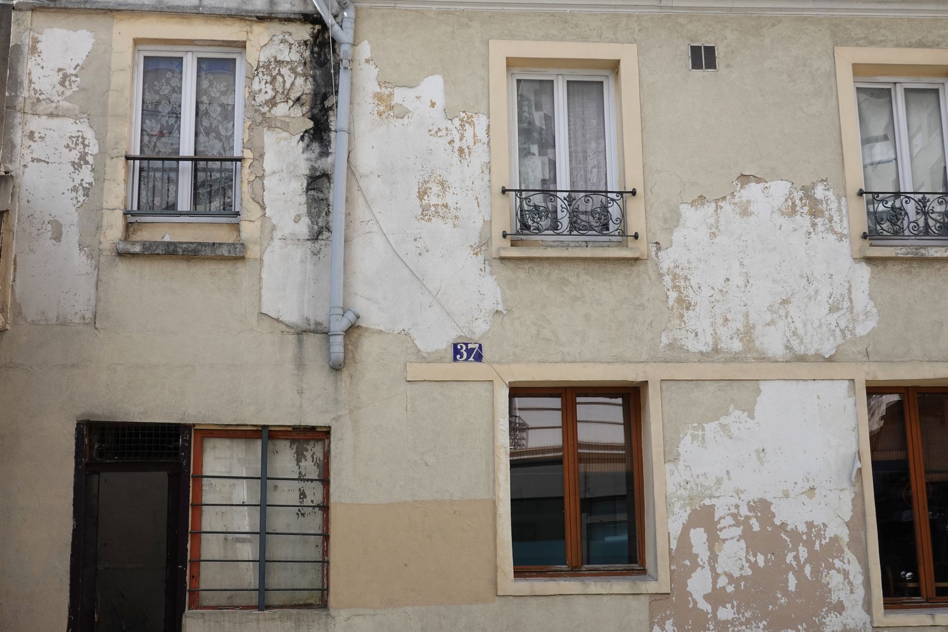 Façade d'appartement délabrée, peinture beige écaillée, trois fenêtres à grilles en fer.