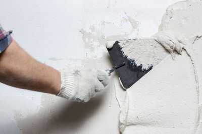 Eine behandschuhte Hand verteilt mit einer Kelle weissen Putz auf einer Wand.
