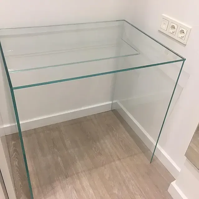 Un pequeño escritorio de vidrio está sobre un piso de madera al lado de una pared.