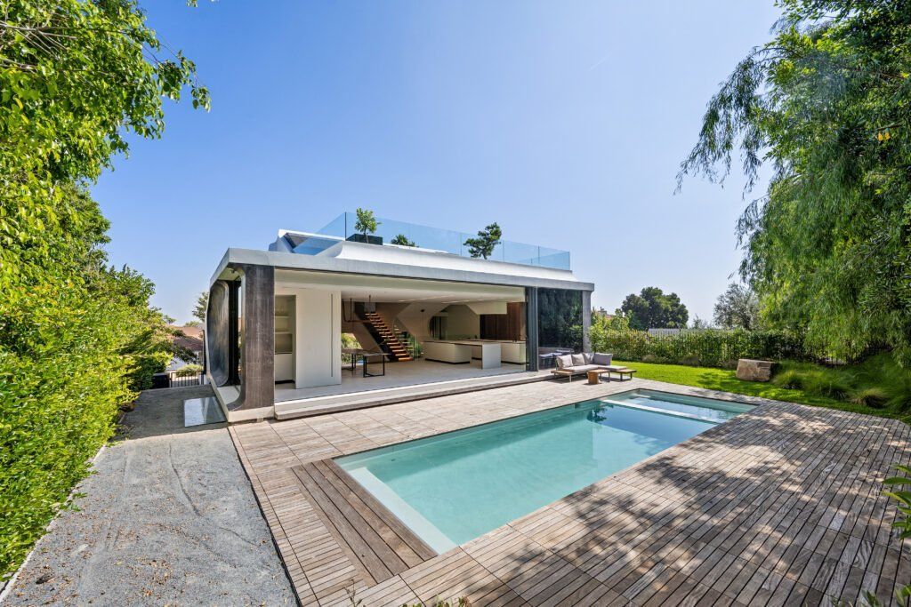 Maison moderne avec piscine rectangulaire, terrasse en bois et végétation luxuriante sous un ciel bleu azur.