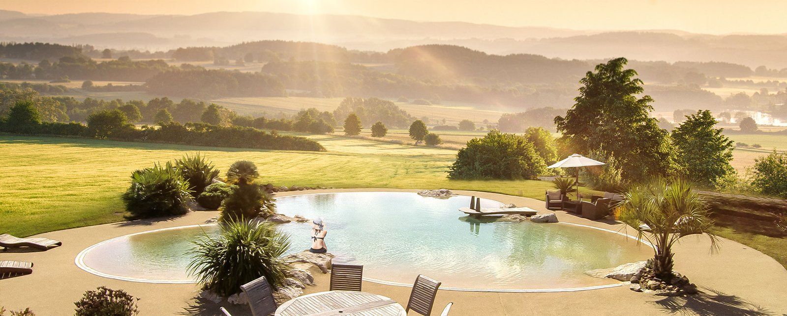 Vue panoramique sur une piscine naturelle et une terrasse avec un paysage montagneux au coucher du soleil.