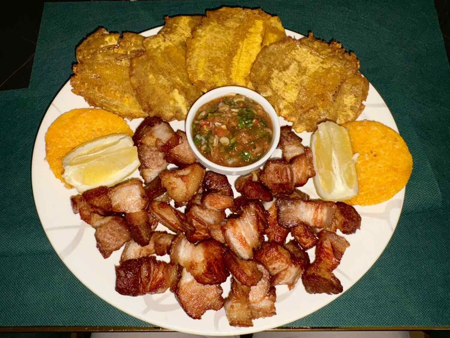 Plato de panceta de cerdo frita, tostones y salsa para mojar, con gajos de limón.