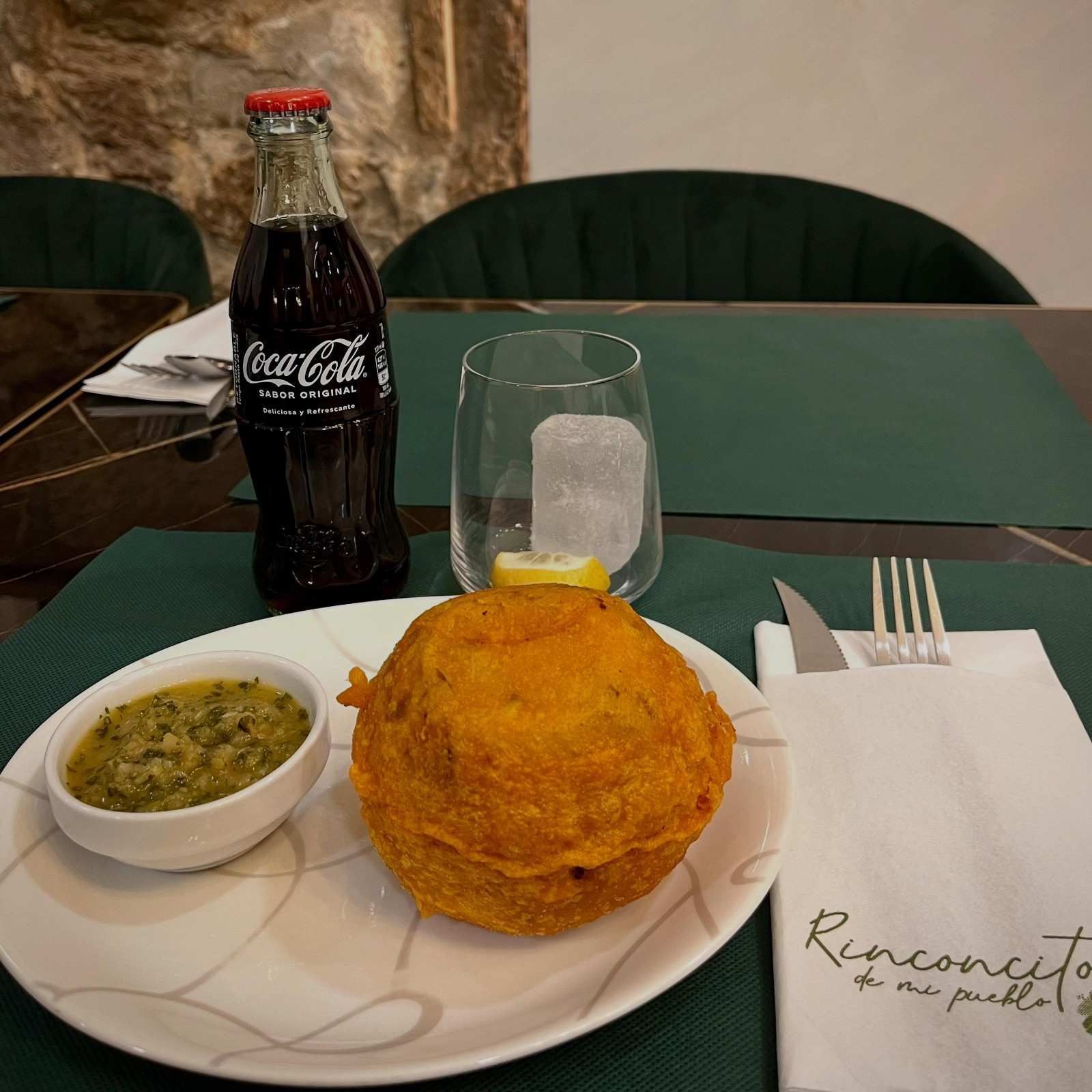 Plato con comida frita y salsa, un vaso, una botella de Coca-Cola y cubiertos sobre una mesa verde.