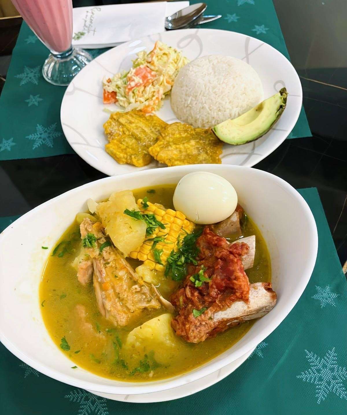 Plato con comida: sopa con carne, maíz y huevo. Plato aparte con arroz, plátanos, ensalada y aguacate.