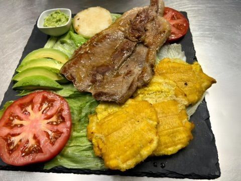Plato de bistec con tostones, rodajas de aguacate, tomate, guacamole y arepa en bandeja negra.