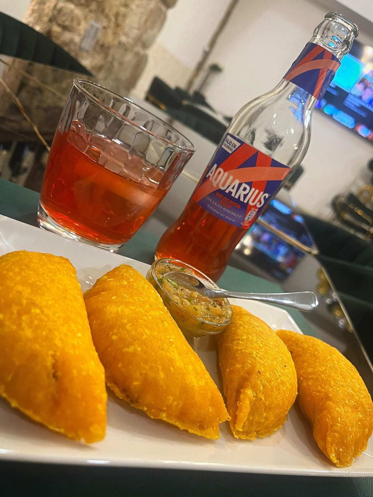 Plato de empanadas fritas, copa de bebida roja, botella de Acuario y mostaza.