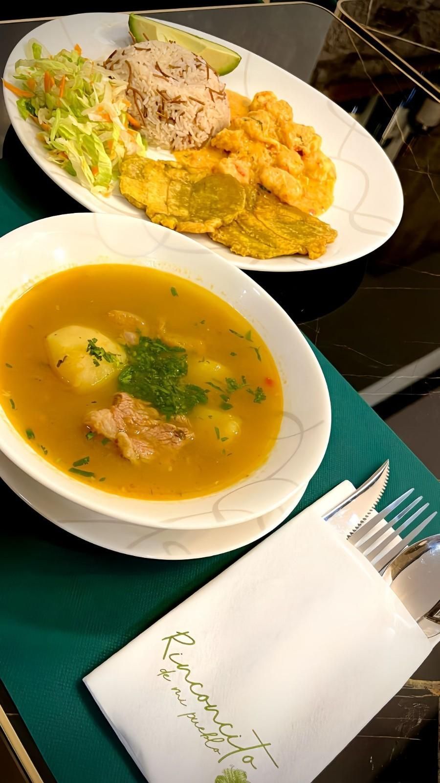 Sopa y plato de arroz, frijoles y plátanos, con cubiertos y servilleta sobre mantel verde.