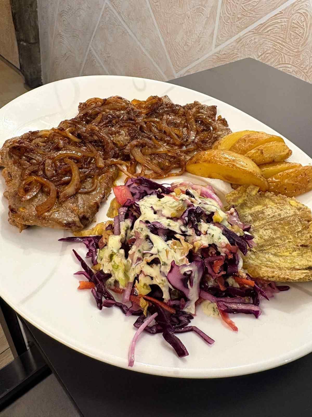 Bistec con cebollas caramelizadas, patatas asadas, ensalada de col y un panqueque de patata frita en un plato blanco.