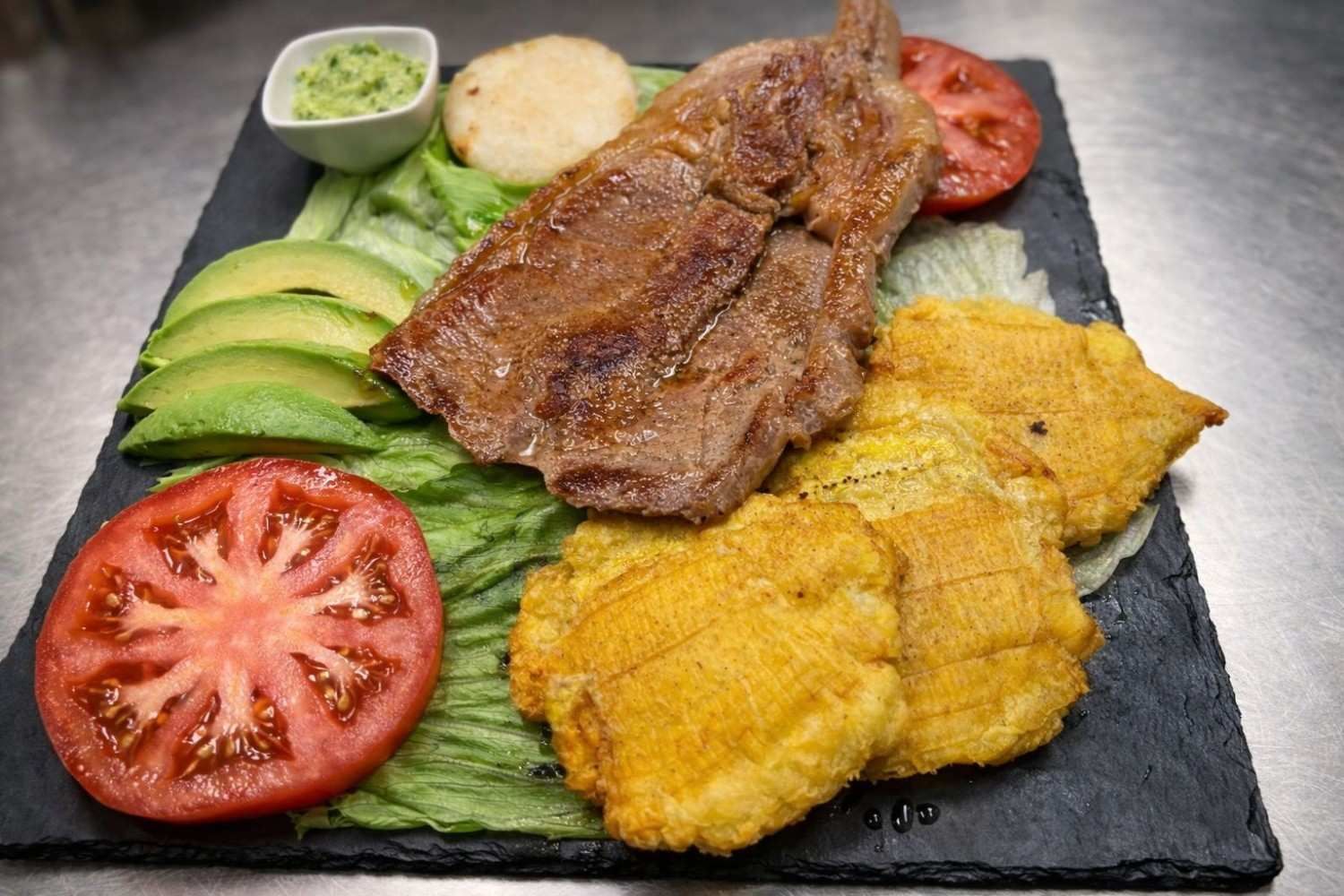 Carne a la parrilla con plátanos fritos, aguacate, tomate, arepa y salsa en plato negro.