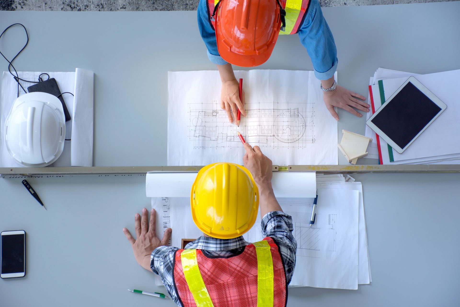Deux ouvriers du bâtiment, casques de chantier sur la tête, examinent des plans à un bureau, munis d'une tablette et d'un téléphone.