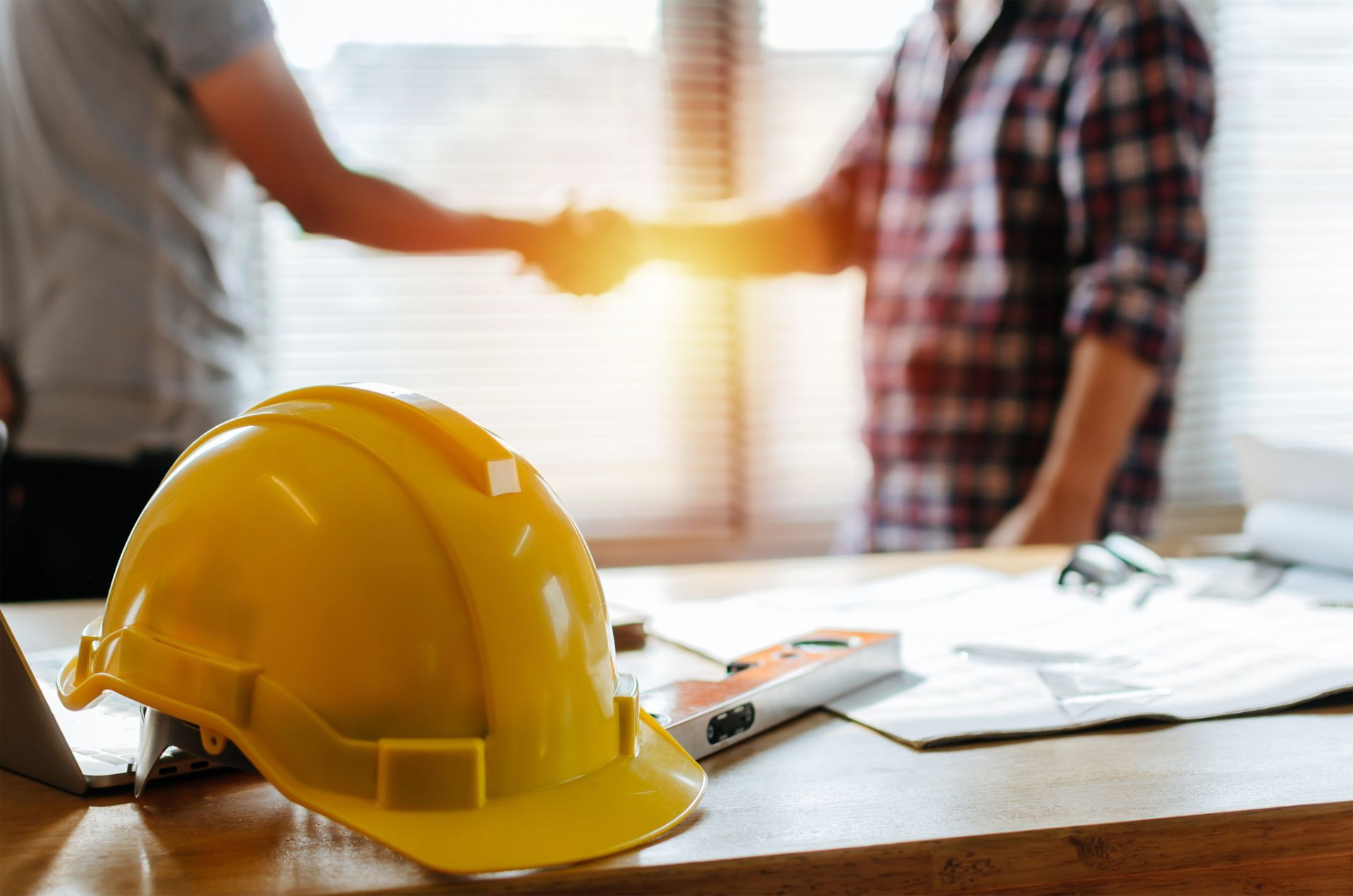 Un casque de chantier jaune posé sur un bureau, avec des personnes se serrant la main en arrière-plan.