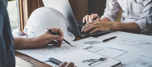 Des mains dessinent des plans sur un bureau, un ordinateur portable et un casque de chantier à proximité, dans un environnement de bureau.