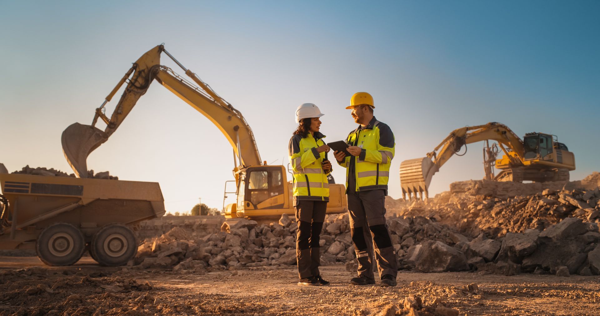 Deux ouvriers du bâtiment en tenue de sécurité se tiennent devant un chantier avec des excavatrices et des camions-bennes derrière eux.