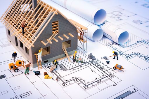 Maison miniature en construction d'après des plans, avec des ouvriers, des outils et des plans architecturaux enroulés.