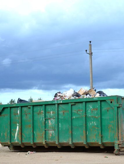 Un gran contenedor verde lleno de basura se encuentra en el medio de un campo de tierra.