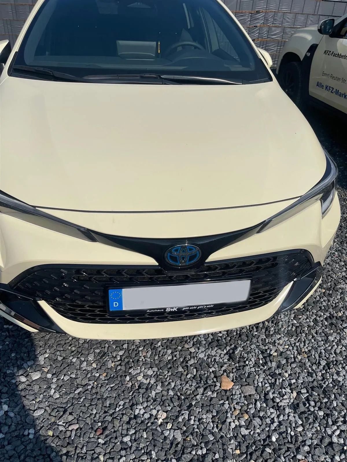 Frontansicht eines weißen Toyota, der auf einem Schotterplatz geparkt ist, mit schwarzem Kühlergrill und Sonnenspiegelungen.