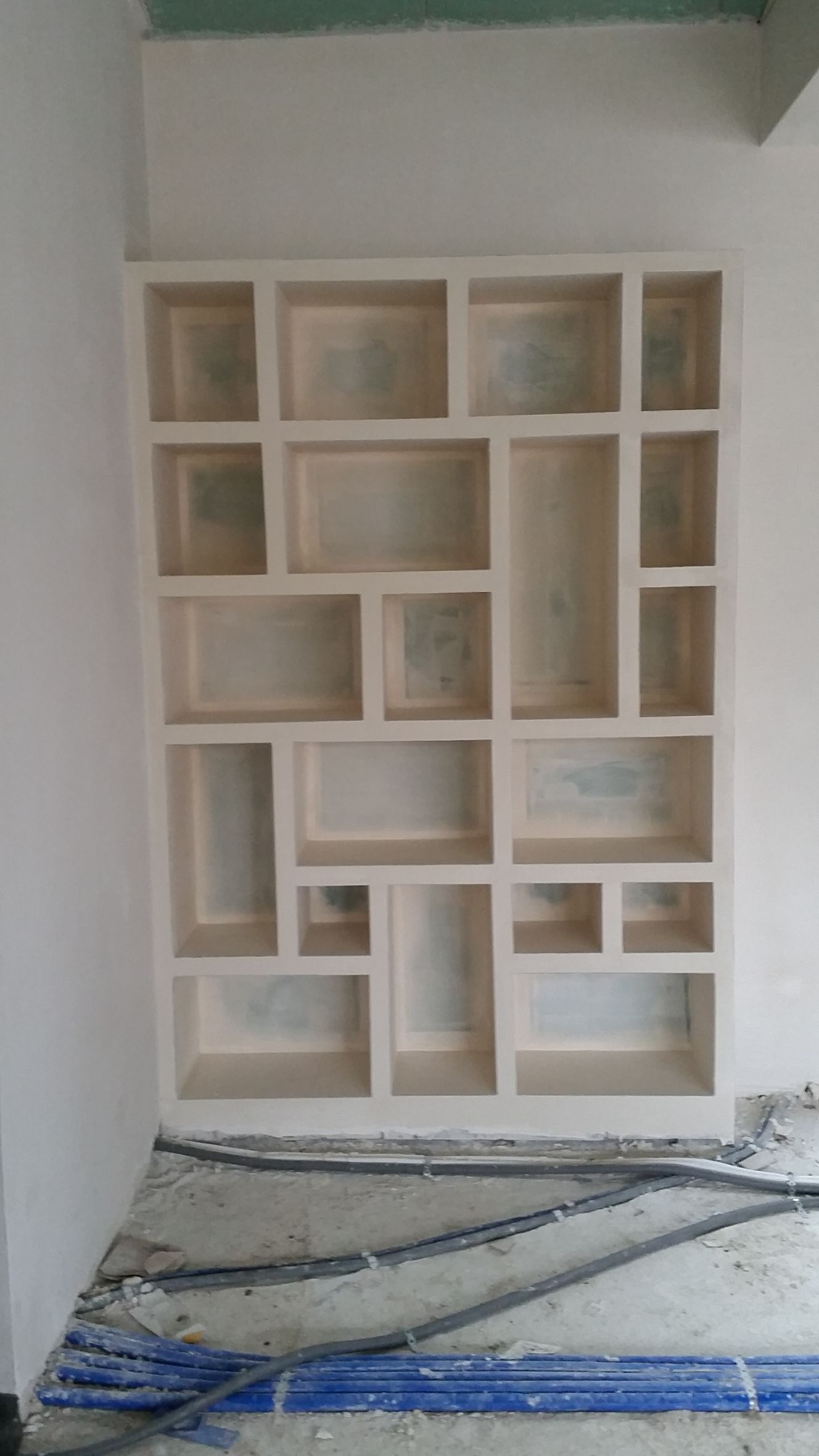 Étagère murale géométrique vide dans une pièce en travaux, avec sol recouvert de plastique et murs non finis.