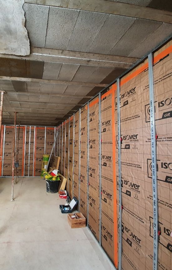 Zone de construction intérieure avec panneaux muraux isolés, ossature orange et outils le long d'un étroit couloir en béton.