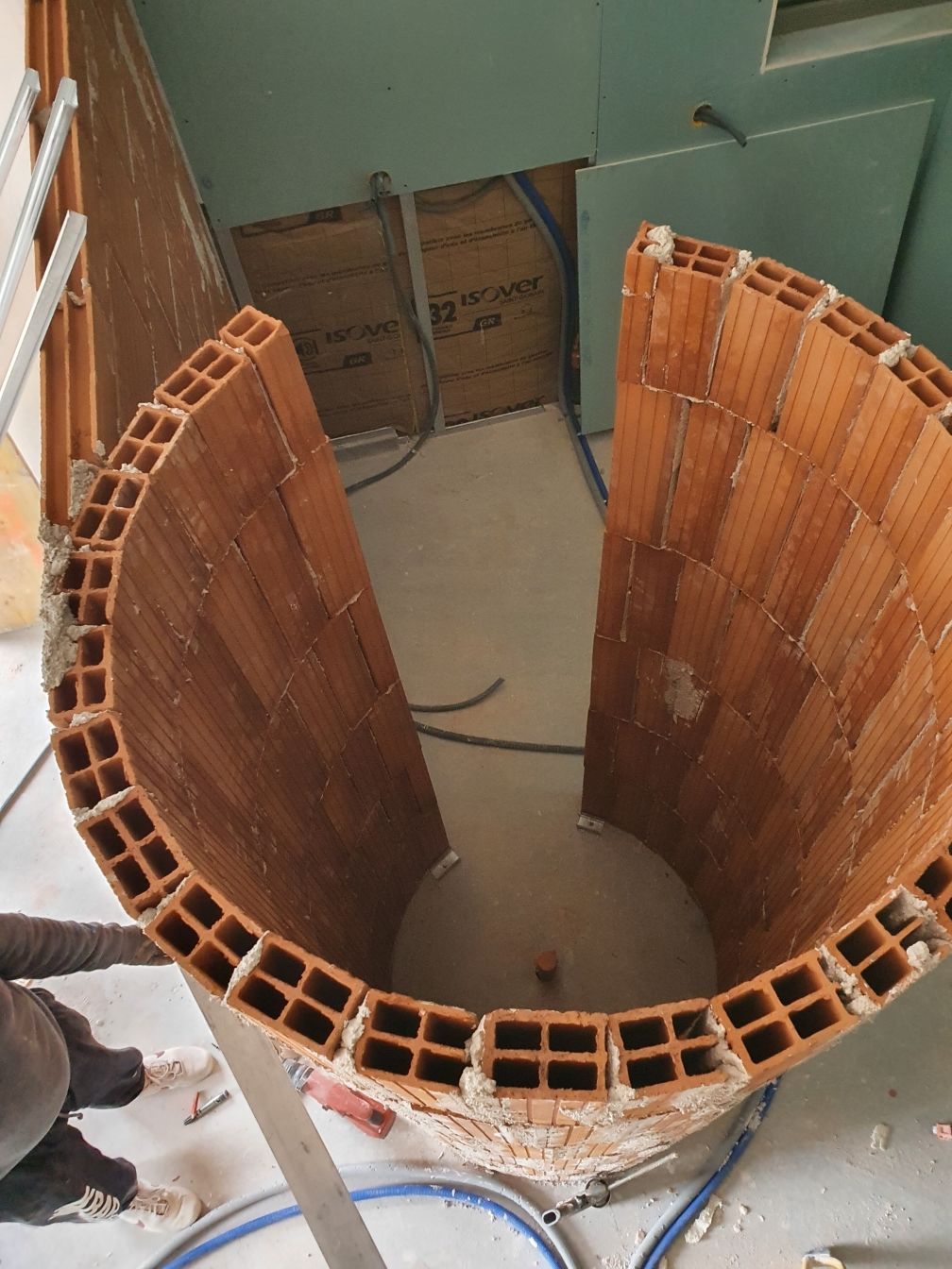 Mur incurvé en briques orange en construction autour d'une ouverture centrale dans une pièce jonchée de débris au sol.