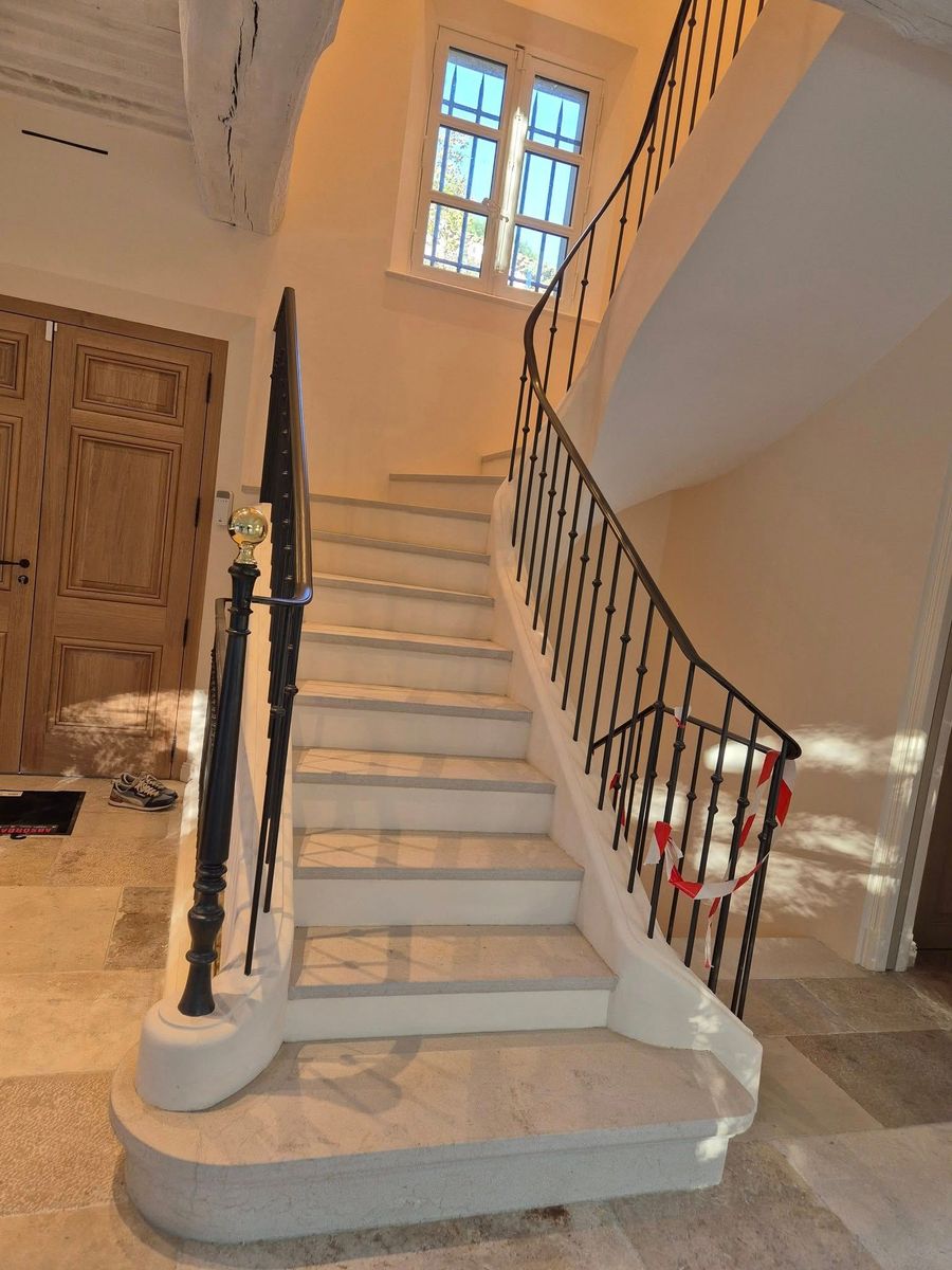 Escalier intérieur avec marches blanches, rampe foncée et palier éclairé par une fenêtre dans un couloir beige.